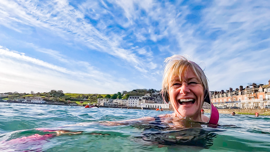 A white woman in her fifties beams. She is swimming in the sea at Swanage Bay which is visible behind her. The sun is shining.