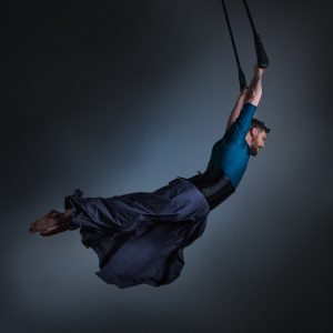 Joel - a built male presenting person hangs from a black trapeze. There is a feeling of motion to the picture as the trapeze looks like its being pushed to the right of the image, and Joel's body is arched underneath it, pointed toes reaching to the opposite side. In this picture they have a short back and sides haircut, and a trimmed beard. There is a blue hue to the picture that matches their silky teal button up shirt, with a black corset over the shirt, and a blue grey skirt fluttering around their ankles.