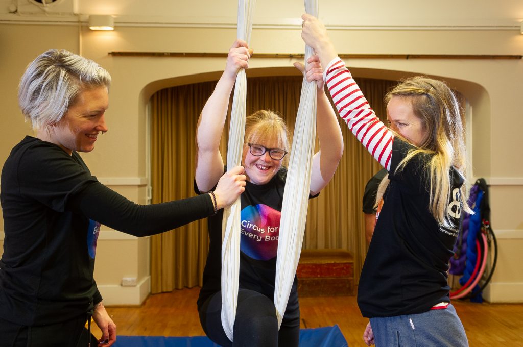 A member of Extraordinary Bodies Young Artists smiles as she is assisted into the silks and swings on them.