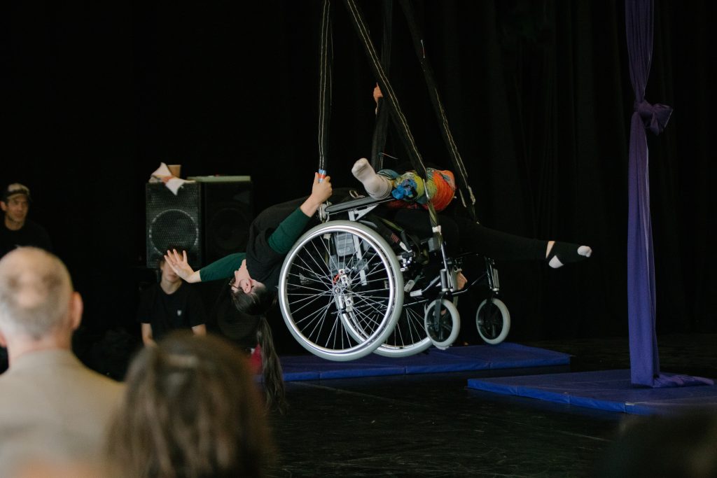 An artist stretches backwards from their wheelchair. They are suspended in an aerial rig and look elegant.