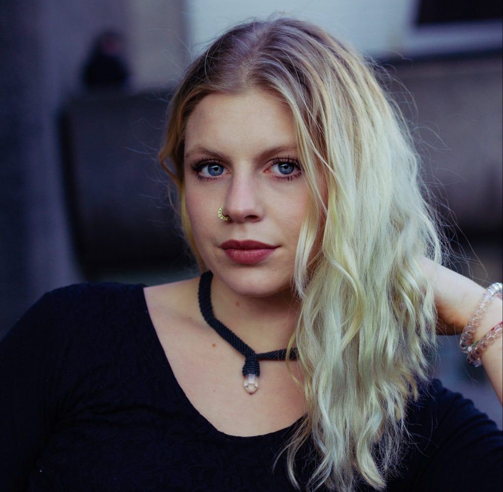 A white young woman with fair hair, dressed in a black top, and wearing a bold necklace.