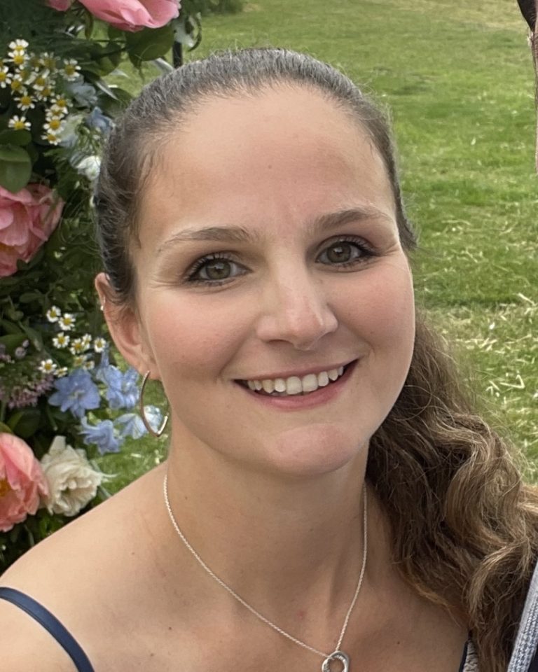 A white woman with curly dark hair, in a slicked back ponytail. She is in a garden with flowers behind her.