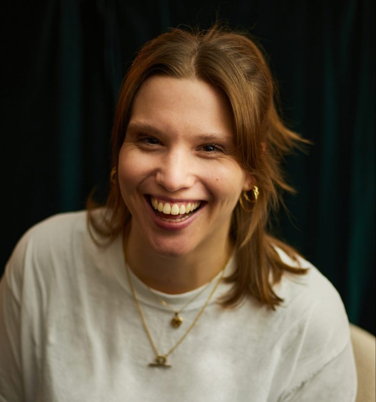 A white woman with shoulder length fair hair smiles at the camera. She wears a white top and two gold necklaces.