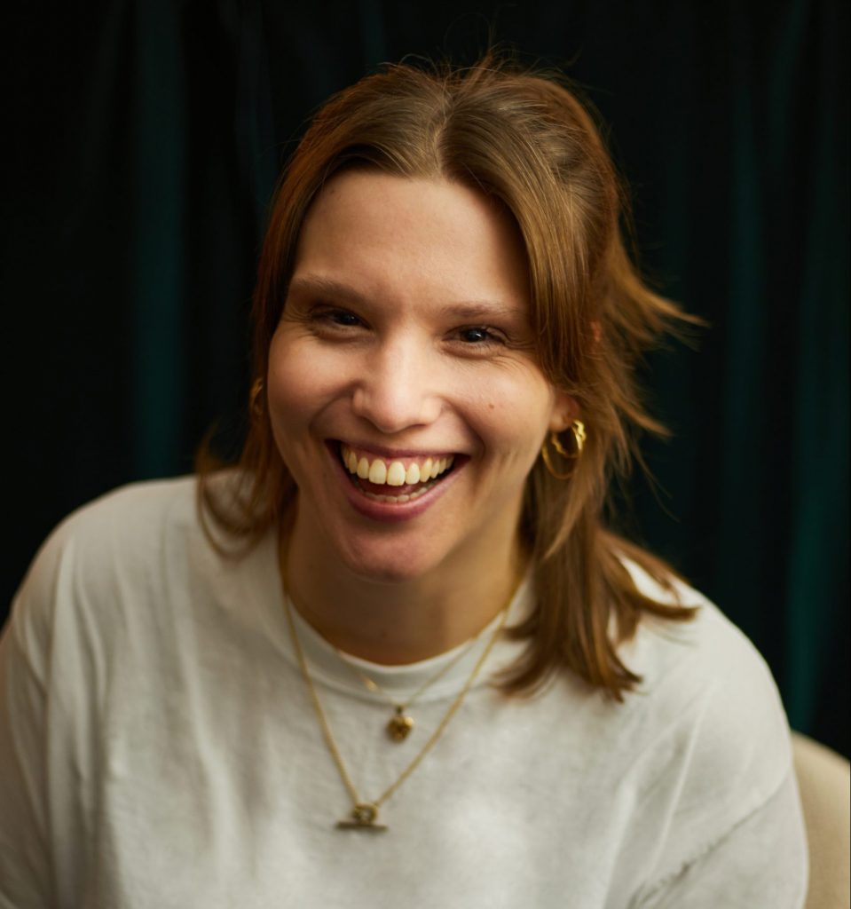 A white woman with shoulder length fair hair smiles at the camera. She wears a white top and two gold necklaces.