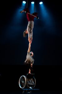 Tilly Lee-Kronick and Jonny Leitch perform aerial work. Tilly hangs upside down from a trapese, holding Jonny by his wrist - he suspends his wheelchair from his other hand as as they take flight.