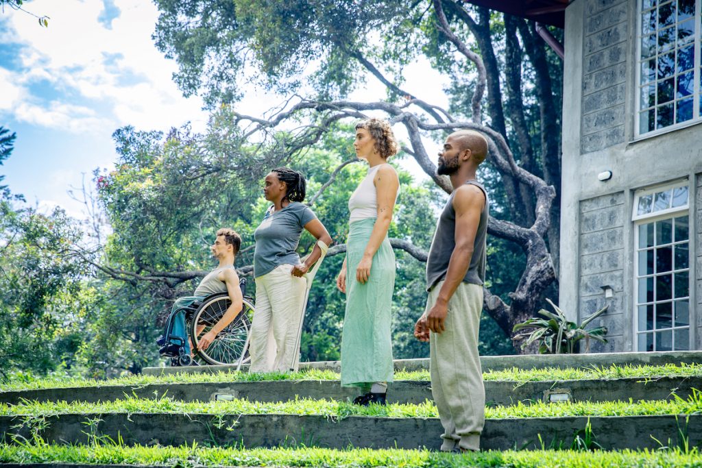 The four dancers stand in a terraced garden on a sunny day with a grand, Victorian building in the background. On the left, on the top tier of the terracing, is Johnny who sits in his wheelchair. Next is Beatrice, who uses crutches to stand. On the next level down is Tilly. On the lowest tier, closest to the camera, is Chris. All four gaze off camera to the left. They all look strong and purposeful.