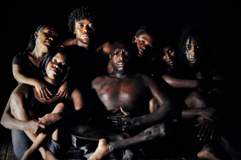 A group of seven Kenyan dancers sit tightly grouped. The men are shirtless, and the women wear black. They are grouped in front of a dark background.