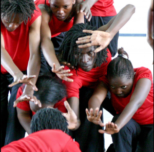 Pamoja Dance Company mid-performance. They are wearing red t-shirts and black trousers. They thrust their hands away from their bodies.