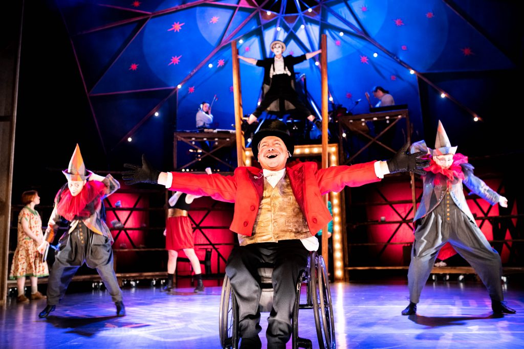 Garry Robson in character as ringmaster Waldo, in Waldo's Circus of Magic and Terror. He wears a black top hat, a red jacket and a hold waistcoat. He throws his arms out wide - the master showman.