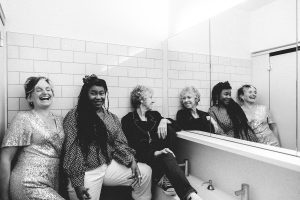 Black and white image - Claire, Jacqui and Karen are in a public bathroom. Karen sits on the edge of the sink and looks in the mirror. Jacqui leans again the wall. Claire is closest to the camera - she's laughing.