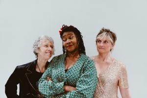 Karen, Jacqui and Claire stand in front of a white background. Karen and Jacqui share a knowing smile, Claire peers towards them, a little suspicious.