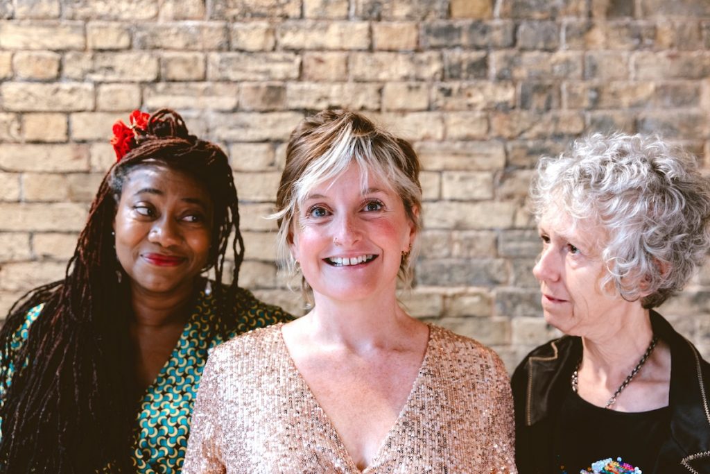 Three women in their mid life stand in front of a brick wall. On the left is Jacqui, a black woman with long brown braids, some of which are swept back and pinned with a red flower. In the centre is Claire, a white woman with short fair hair. The pieces around her face are bleached platinum, and she's wearing a sequinned jumpsuit. She beams at the camera. On the right is Karen, a white woman with curly grey hair. She wears a pride t-shirt under a black leather jacket, and is looking at Claire with disbelief.