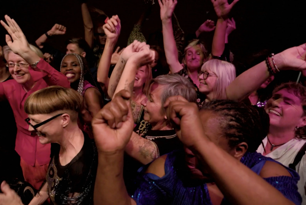 #DanceThroughMidLife A diverse group of women in their mid life dance. They throw their arms up, smiling - disco lights colour the image.