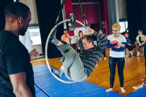 A young man with Down syndrome balances on an aerial hoop in a gym. A supportive instructor stands nearby, observing with encouragement.