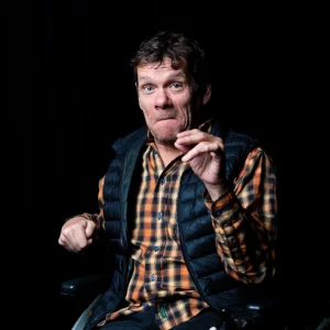 Jamie Beddard looking straight at the camera, with a black background. He's wearing a checked shirt, and holding one hand up.