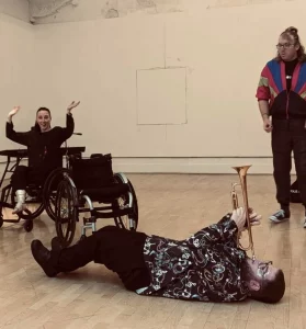 Harry Bassett during a workshop with two other people in the room. Harry is lying on the floor on his back and playing the trumpet.