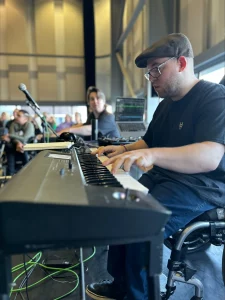 Harry Bassett playing keyboard during a performance. You can see another performer in the background and an audience.